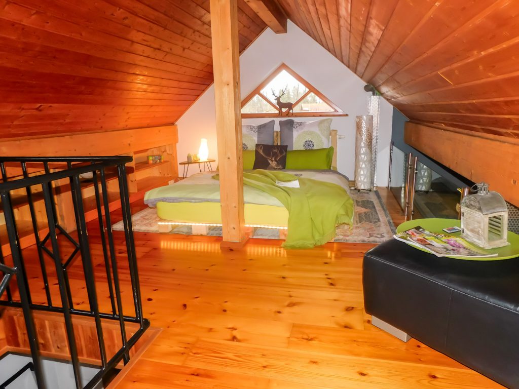 Schlafzimmer unterm Dach mit einem Doppelbett, Beleuchtung und einem Beistelltisch. Links sind außerdem Regale zu sehen.